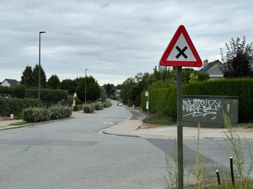 Triangle à bord rouge portant une croix noire, planté avant un carrefour de rue résidentielle